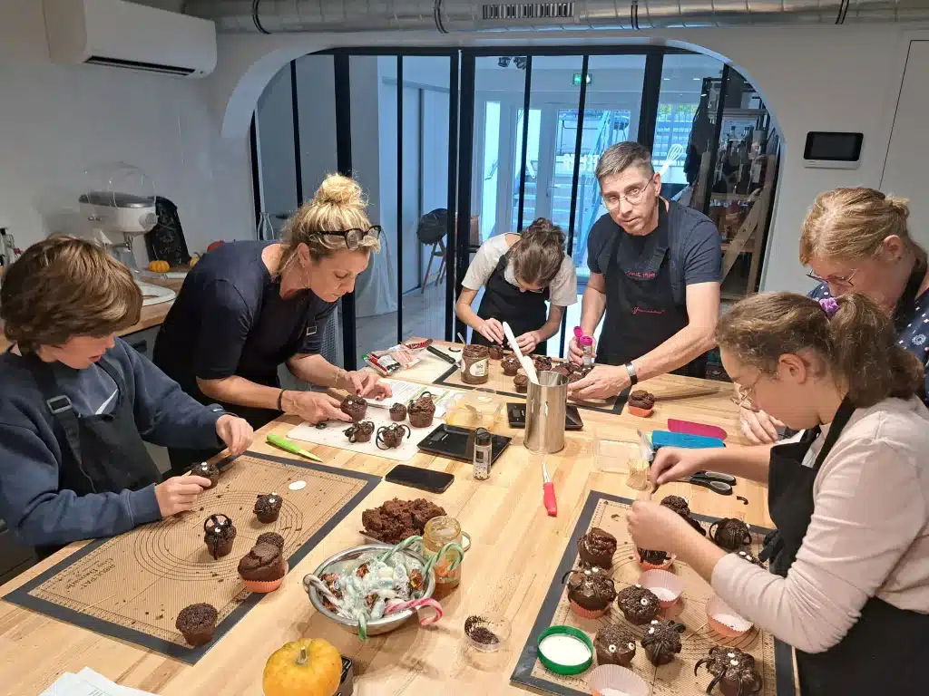 Un groupe de personnes, composé d'enfants et d'adultes, décore ensemble des petits gâteaux au chocolat à une grande table d'atelier cuisine et pâtisserie, concentrés sur leurs tâches et entourés d'outils et d'ingrédients de boulangerie professionnels.