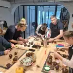 Un groupe de personnes, composé d'enfants et d'adultes, décore ensemble des petits gâteaux au chocolat à une grande table d'atelier cuisine et pâtisserie, concentrés sur leurs tâches et entourés d'outils et d'ingrédients de boulangerie professionnels.
