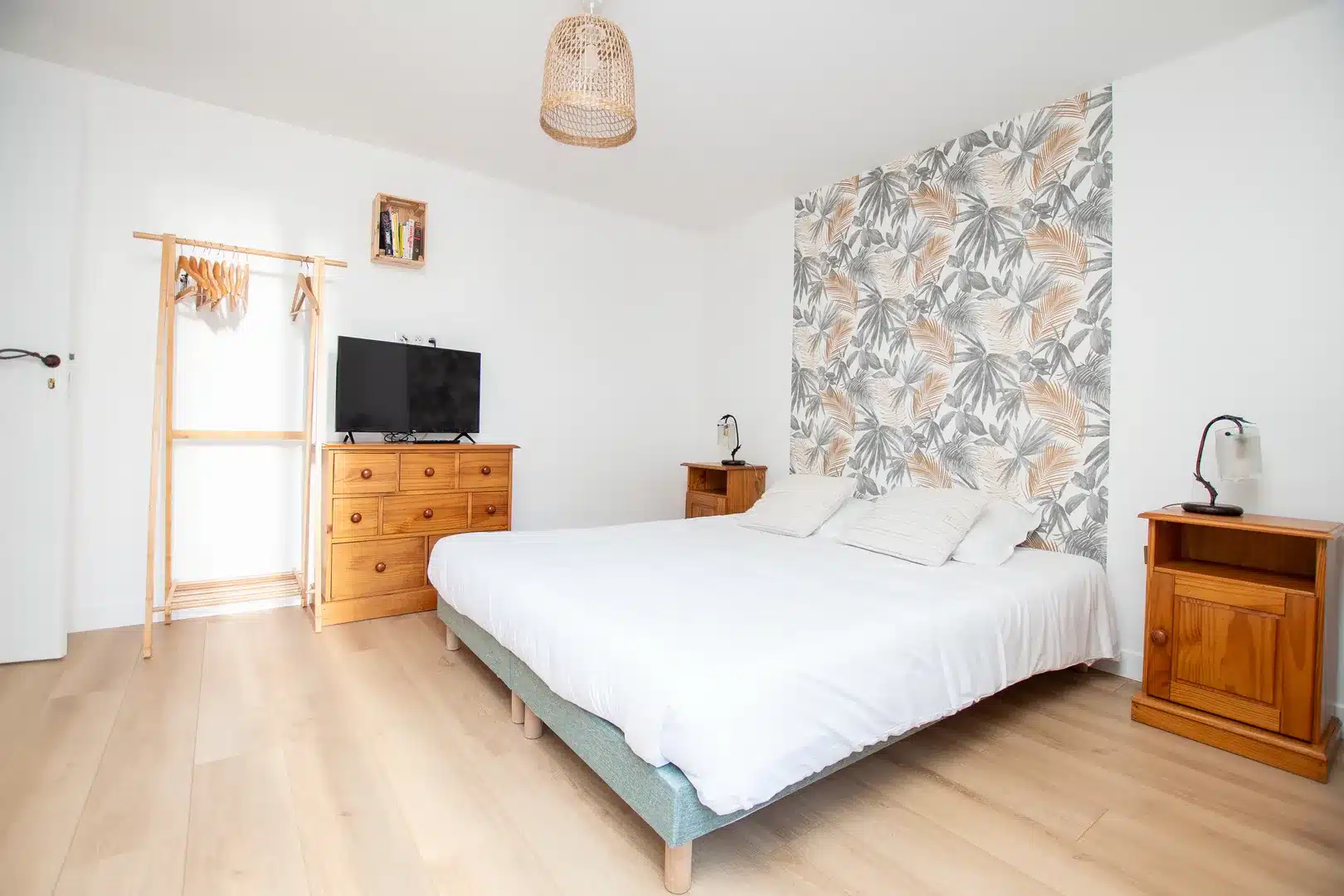 Chambre d'hôte lumineuse avec un lit double, une literie blanche, des tables de nuit en bois, une commode avec une télévision, un portemanteau près de la fenêtre et un mur avec un papier peint de feuilles tropicales. Un sol en bois clair et un plafonnier tressé complètent l'espace.