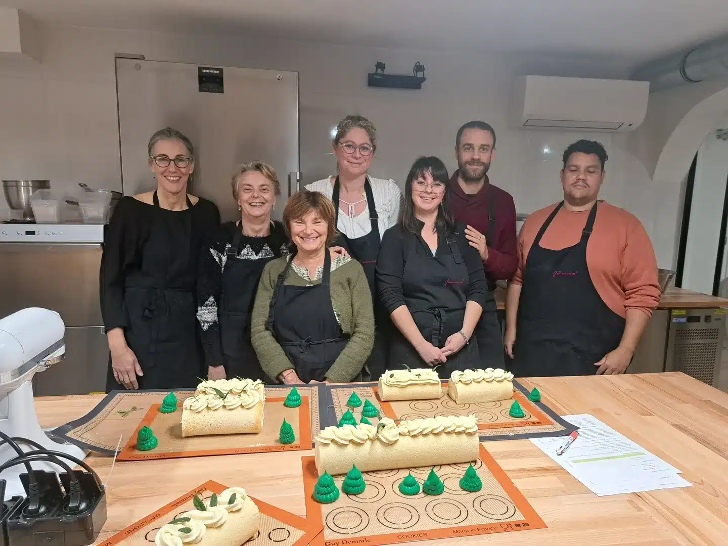 Six personnes en tablier se tiennent debout et sourient derrière un comptoir où se trouvent quatre gâteaux décorés en forme de bûche de Noël et des décorations vertes en forme de sapin, dans un atelier de cuisine et de pâtisserie. Des outils de cuisson et une feuille de recettes sont également visibles sur le comptoir.