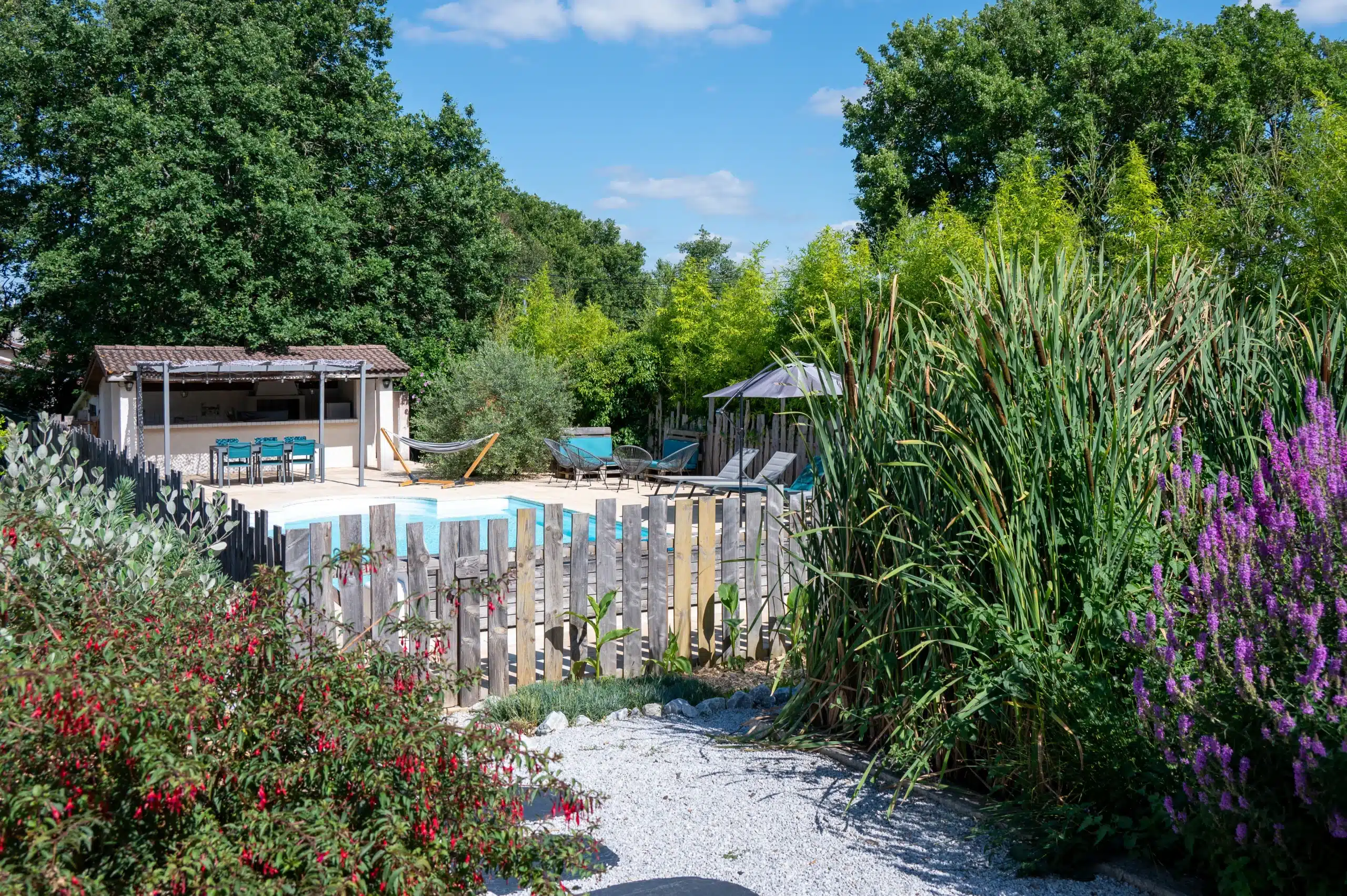 Une piscine extérieure clôturée, entourée d'une végétation luxuriante, de plantes à fleurs, de chaises longues et d'un coin salon ombragé sous un kiosque, vous attend dans ces charmants gîtes et chambres d'hôtes, avec un ciel bleu clair au-dessus de la tête.