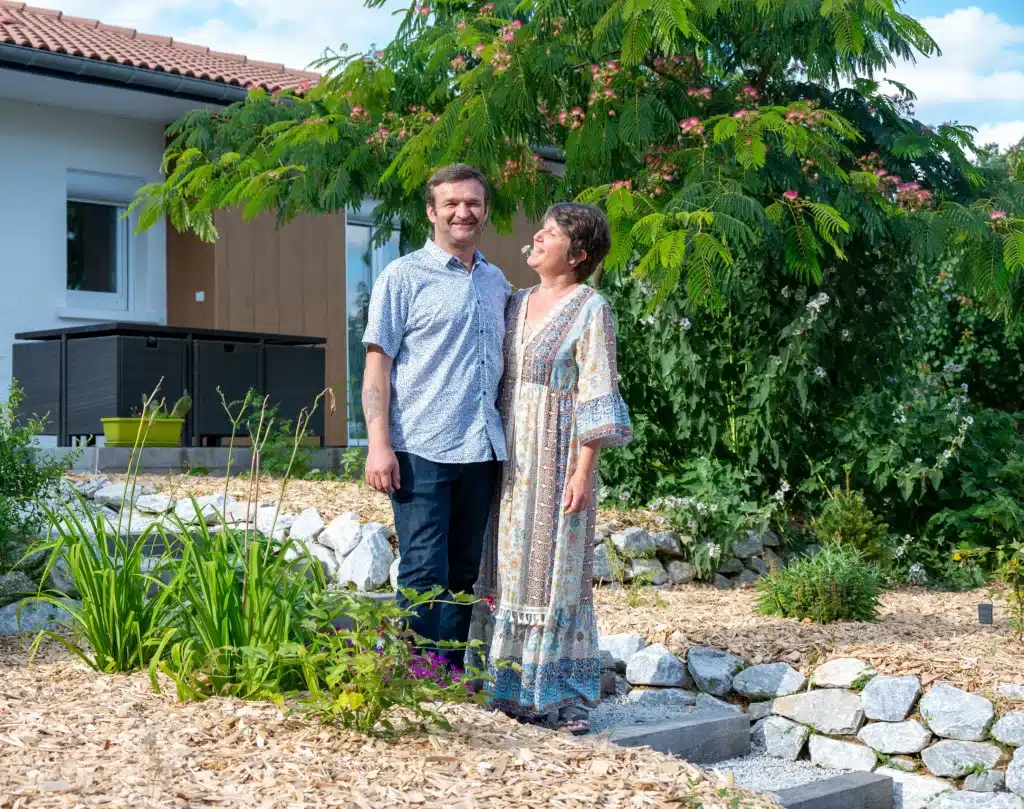 Les gérants de l'Escapade Nature et Gourmande sourient dans un jardin, entourés de verdure et de marches en pierre, avec une maison blanche et des arbres en arrière-plan, par une journée ensoleillée.