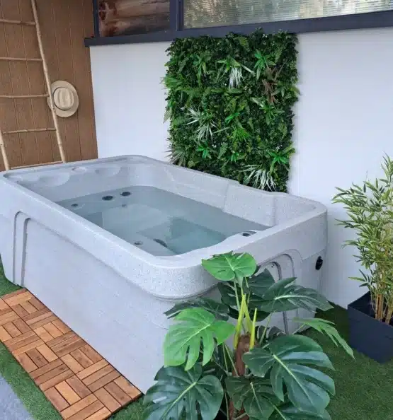 Un jacuzzi extérieur moderne rempli d'eau est entouré de plantes en pot, d'un mur vertical de plantes vertes et d'une natte en bambou au sol, créant ainsi une atmosphère relaxante, semblable à celle d'un spa.