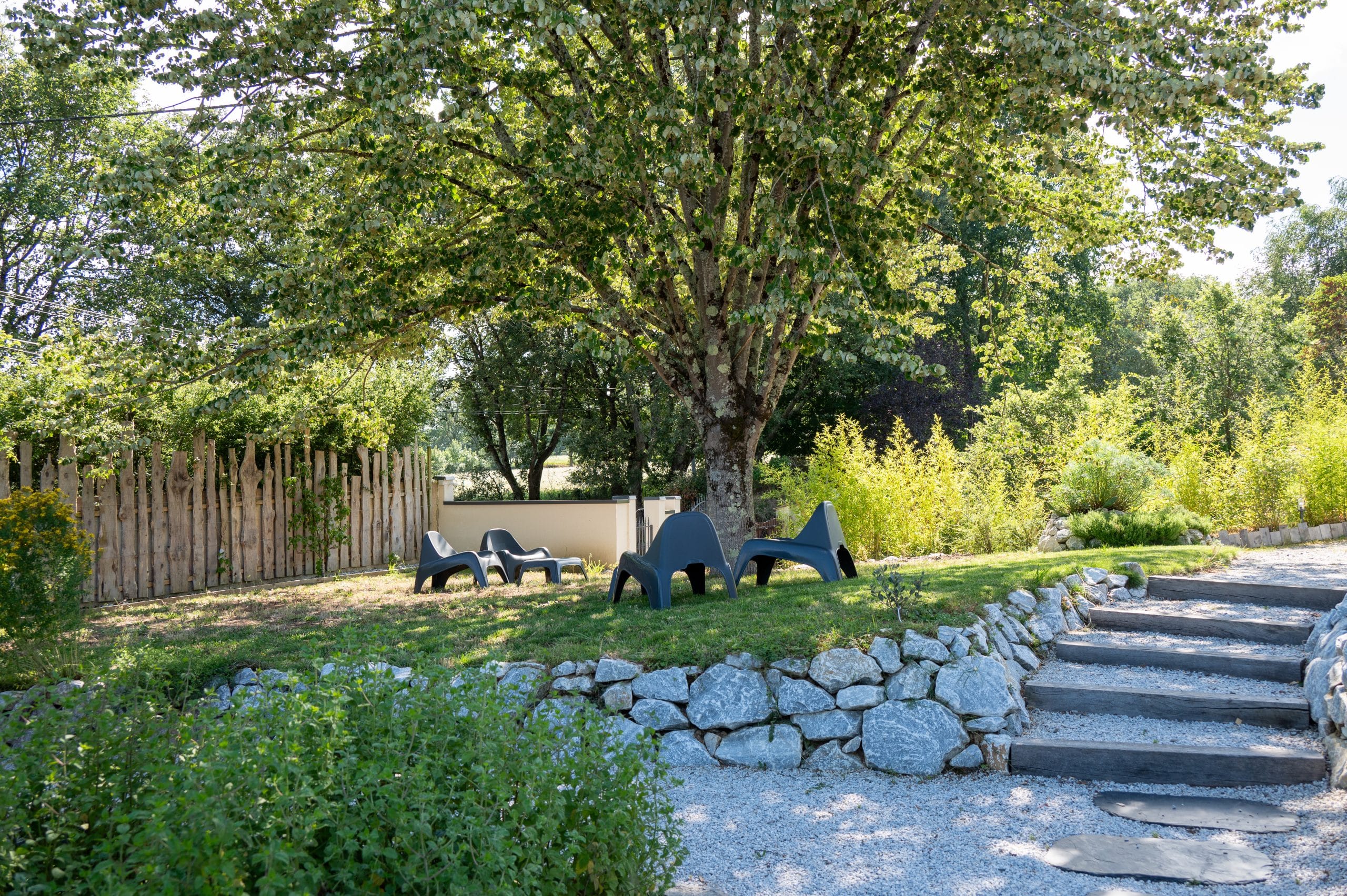 Quatre chaises longues noires sont installées sous un grand arbre feuillu dans un jardin ombragé, bordé d'un mur en pierre et d'une clôture en bois, avec des marches en pierre qui montent à travers la verdure.