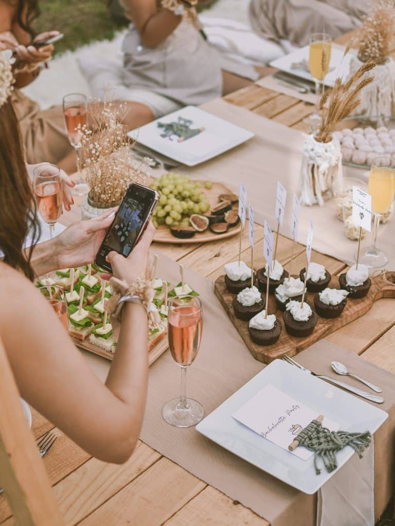 Une femme prend une photo d'une table de pique-nique rustique sur laquelle sont disposés des amuse-gueules, des petits gâteaux surmontés d'un drapeau, des raisins, du vin rosé et une carte de menu décorative, le tout entouré de personnes portant des vêtements décontractés aux couleurs neutres.