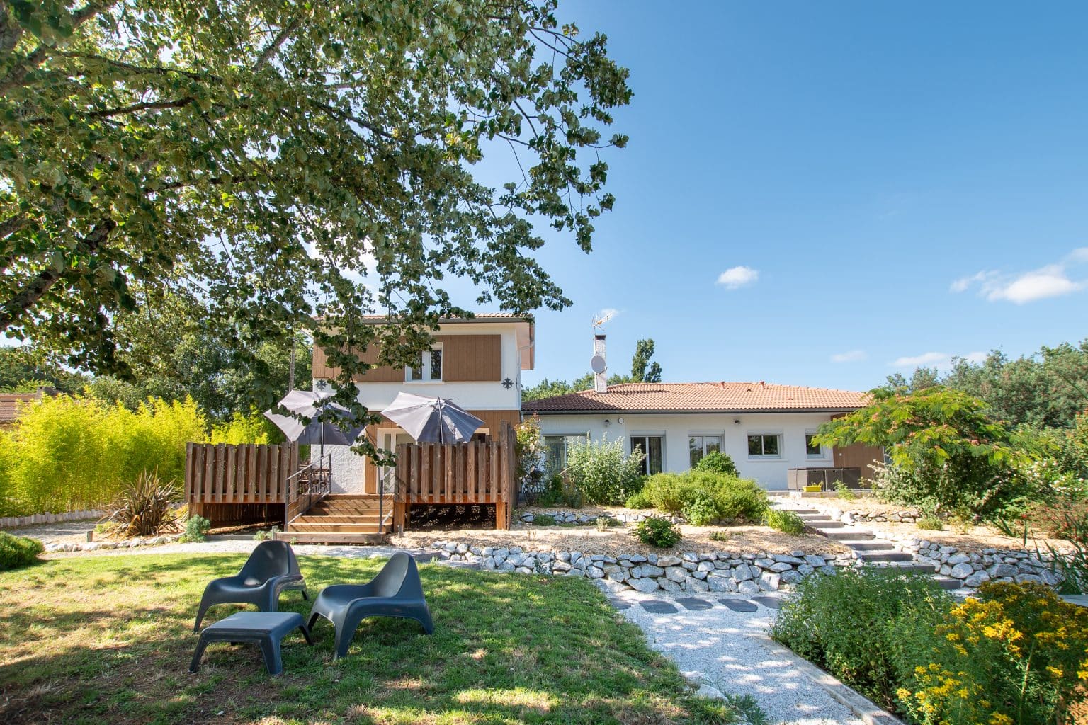Une maison moderne de Gites et chambres d'hôtes avec une terrasse en bois, des parasols extérieurs et un jardin paysager. Deux chaises noires sont assises sur l'herbe sous un grand arbre, entourées de verdure luxuriante et d'un chemin de pierre sous un ciel bleu vif.