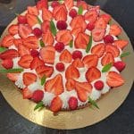 Une tarte ronde garnie de crème fouettée, de fraises coupées en deux, de framboises entières et de petites feuilles de menthe, disposée sur une planche dorée sur fond sombre.