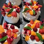 Mini pavlovas garnis de crème fouettée, de fraises, de framboises, de morceaux de fruits orange, de fleurs comestibles et de feuilles vertes, disposés sur un plateau noir.
