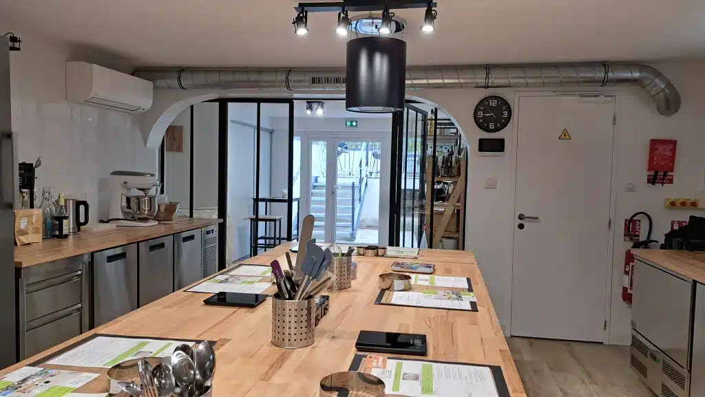 Une salle de classe moderne d'atelier cuisine et pâtisserie avec des postes de travail en bois équipés d'ustensiles, de fiches de recettes et d'outils de cuisine. Des appareils en acier inoxydable et une horloge sont visibles, et des portes en verre s'ouvrent sur un escalier à l'extérieur.
