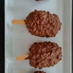 Quatre barres glacées enrobées de chocolat, à la texture croquante et noisetée, sont disposées en rang sur une assiette rectangulaire blanche, chacune sur un bâtonnet en bois.