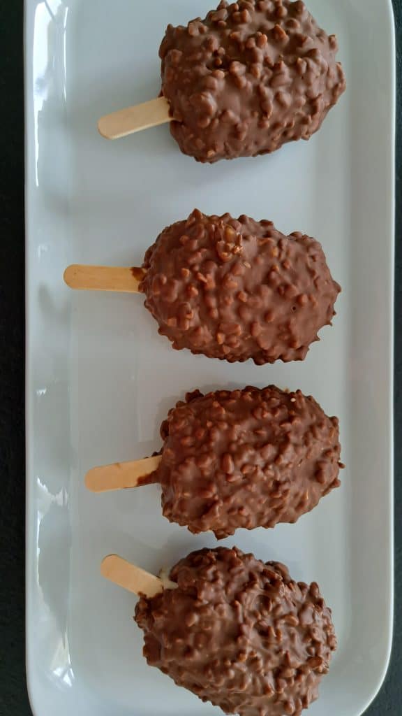 Quatre barres glacées enrobées de chocolat, à la texture croquante et noisetée, sont disposées en rang sur une assiette rectangulaire blanche, chacune sur un bâtonnet en bois.