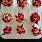 Huit petits desserts garnis de crème fouettée, de fraises en tranches et de feuilles vertes sont disposés proprement sur un plateau blanc. Le fond est sombre et quelques fraises sont placées sur le côté.