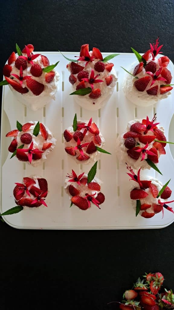 Huit petits desserts garnis de crème fouettée, de fraises en tranches et de feuilles vertes sont disposés proprement sur un plateau blanc. Le fond est sombre et quelques fraises sont placées sur le côté.