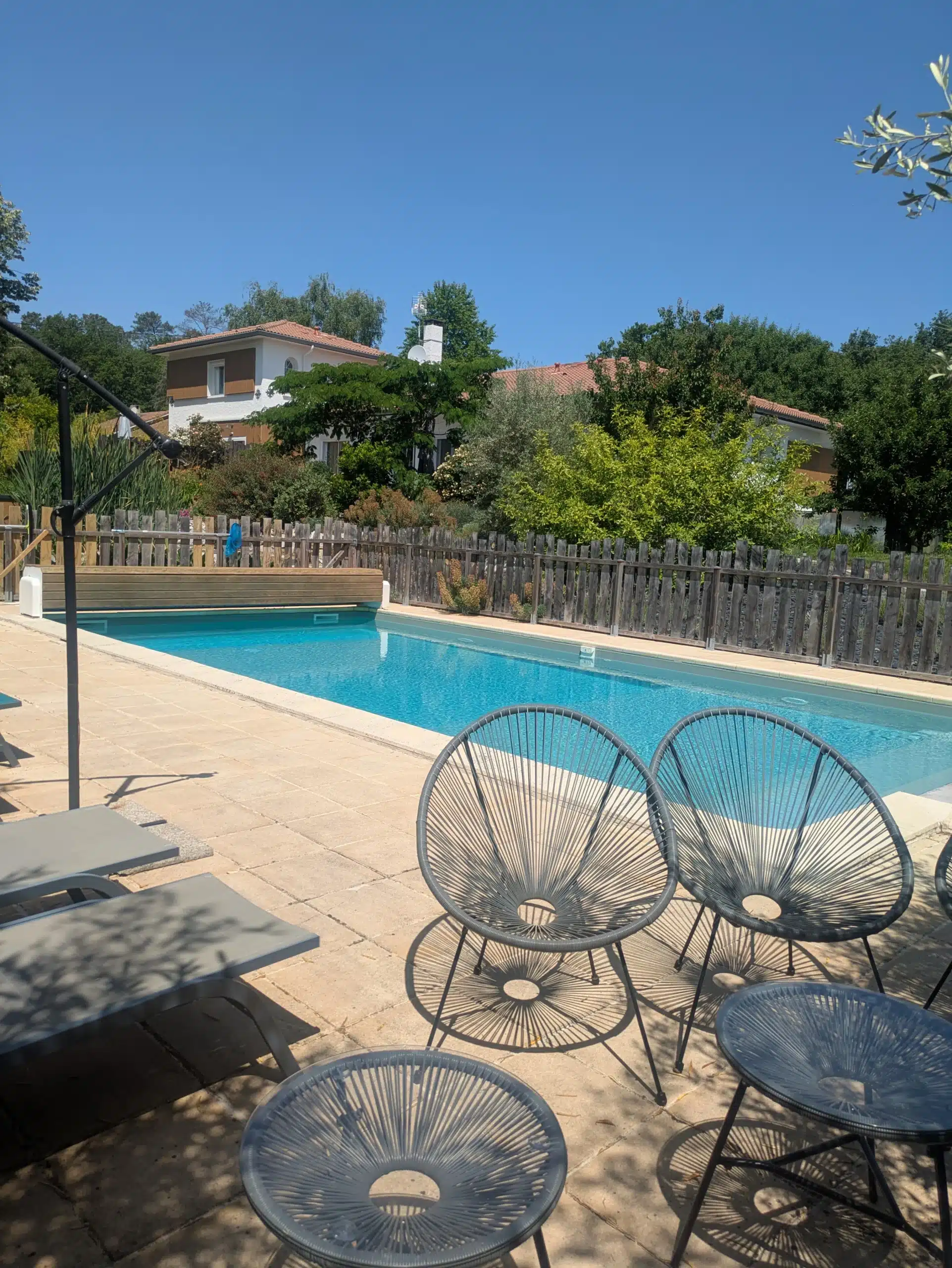 Une piscine extérieure ensoleillée avec des chaises longues et des chaises rondes en fil de fer sur un patio carrelé, parfait pour se détendre dans nos gîtes et chambres d'hôtes, entourés d'une clôture en bois, d'une verdure luxuriante et des maisons voisines sous un ciel bleu clair.