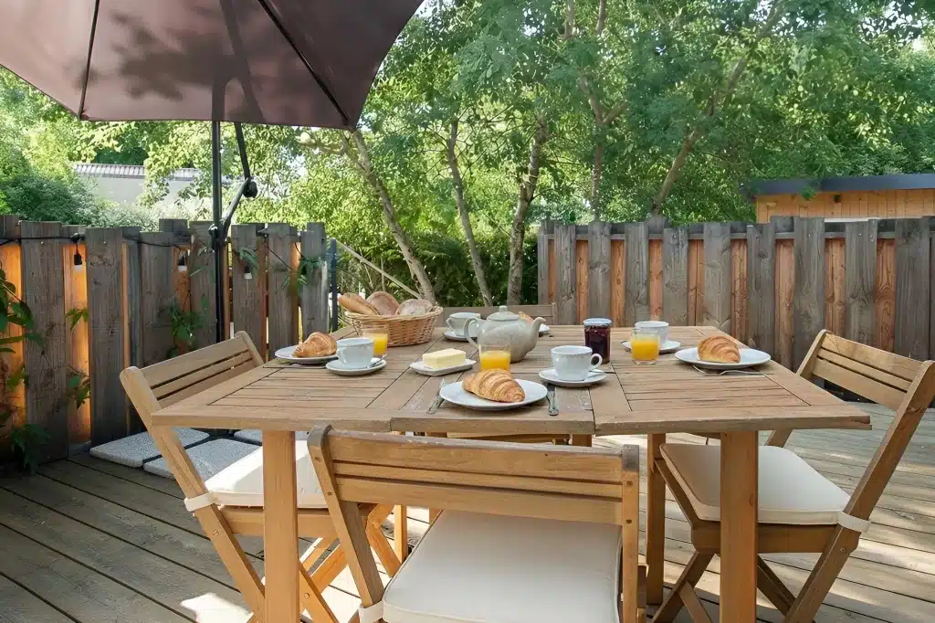 La table en bois du patio d'un Gîtes et chambres d'hôtes est préparée pour le petit déjeuner avec des croissants, du jus, du beurre, de la confiture, une théière et des paniers de pain. Quatre chaises et un grand parasol offrent un confort au milieu des arbres tranquilles de la terrasse.