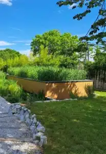 Une jardinière rectangulaire en bois, inspirée du design potager, est remplie de hautes herbes vertes, dans un jardin paysager entouré d'herbe, de pierres et d'arbres, sous un ciel bleu lumineux.