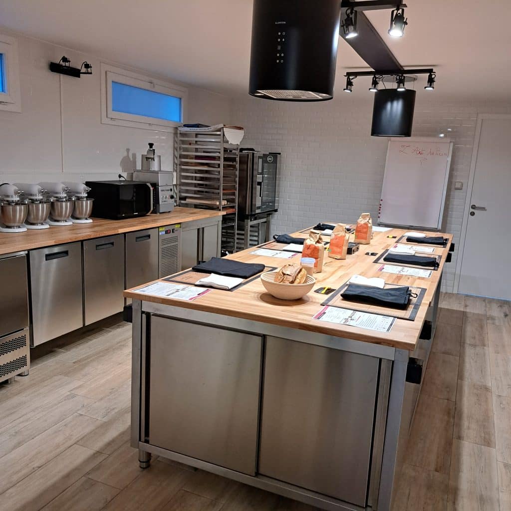 Une salle de classe moderne d'un atelier de cuisine et de pâtisserie avec des comptoirs en acier inoxydable, des mixeurs, des fours et un grand îlot central en bois sur lequel sont disposés des fiches de recettes, des ustensiles et des ingrédients, prêts pour un cours de cuisine. Un tableau blanc se trouve à l'arrière-plan.