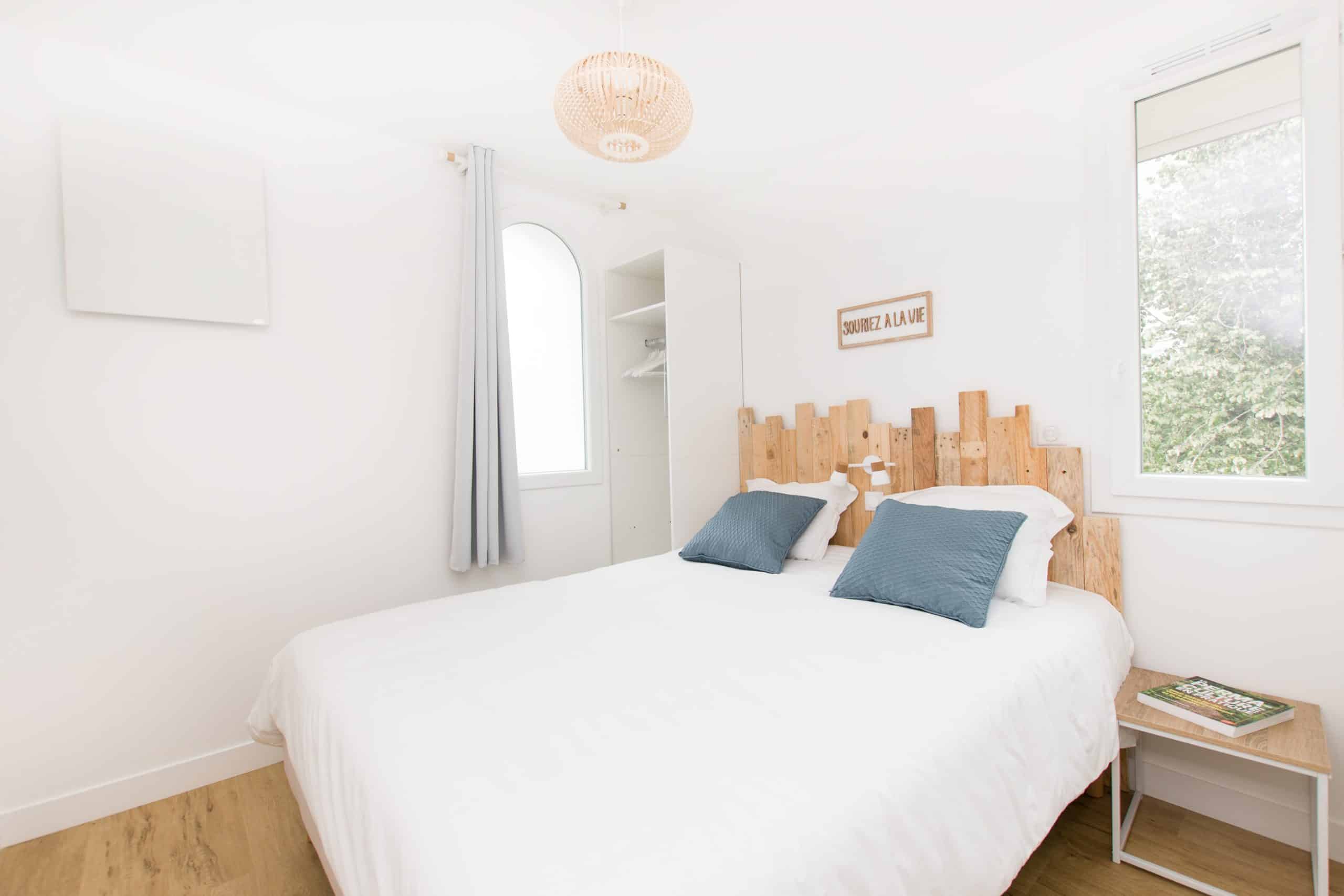 Chambre de gîte lumineuse et minimaliste avec une tête de lit en bois, des draps blancs, deux coussins bleus et une petite table de chevet avec des livres. Plancher en bois clair, murs blancs, grande fenêtre et luminaire suspendu.