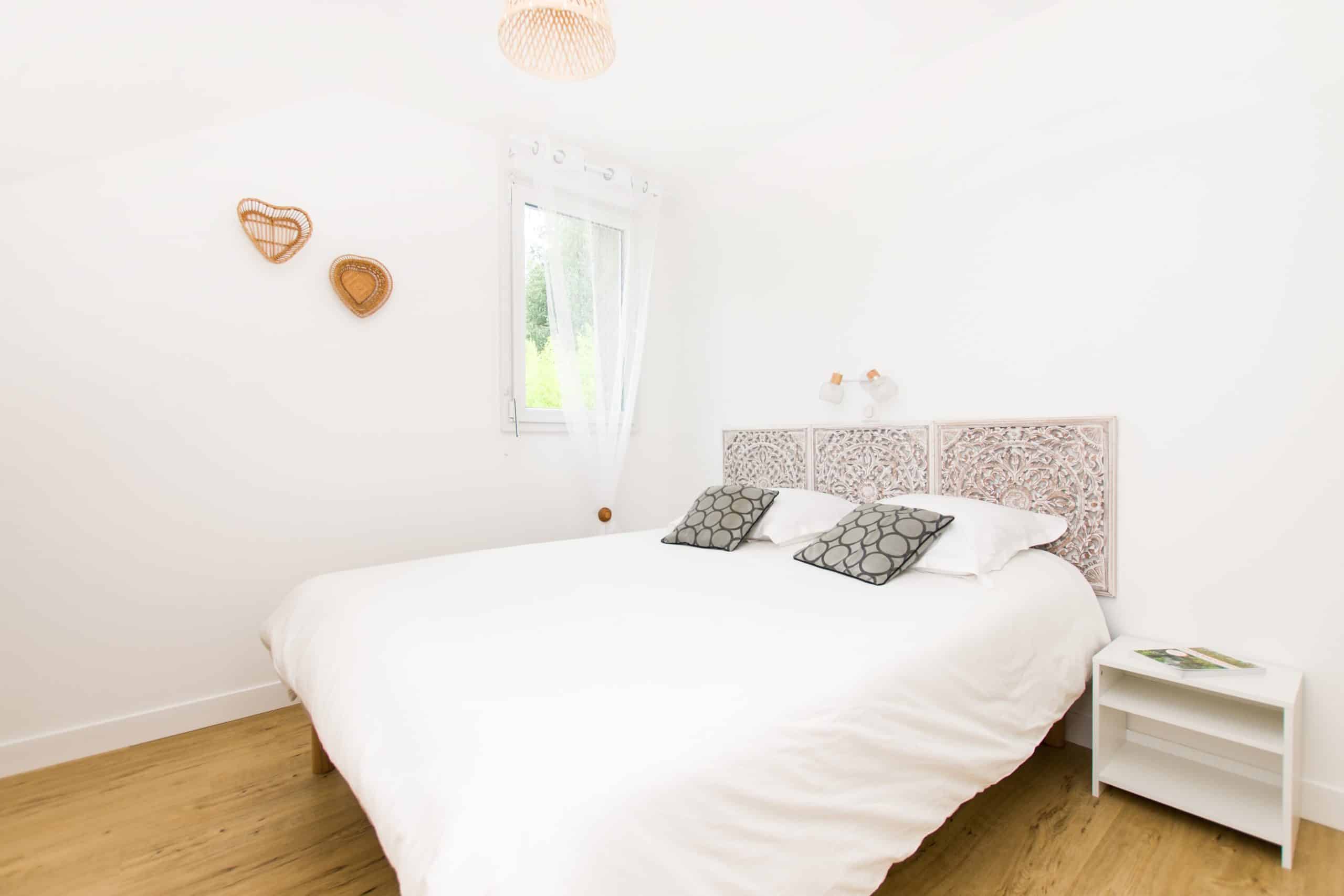 Chambre de gîte lumineuse et minimaliste avec un grand lit blanc, des oreillers à motifs, une tête de lit décorative, une petite table de nuit blanche et un tableau mural en osier ; le sol en bois clair et une fenêtre avec des rideaux diaphanes laissent entrer la lumière naturelle.