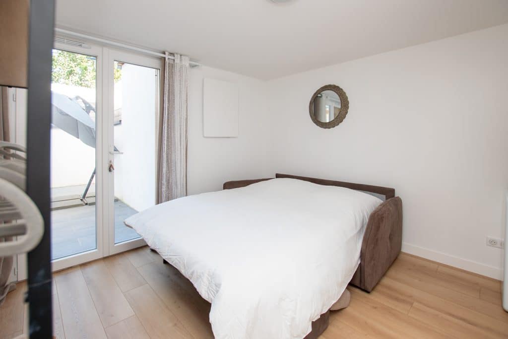 Une petite chambre d'hôte lumineuse avec un canapé convertible recouvert d'une couette blanche, un miroir rond sur le mur blanc et des portes vitrées s'ouvrant sur un patio avec des meubles d'extérieur. Un parquet en bois clair complète la pièce.