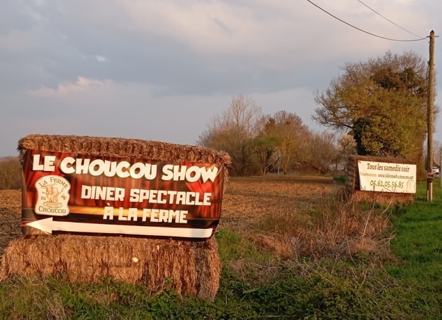 Au bord d'un champ, une grande botte de foin affiche une banderole rouge indiquant Le Choucou Show, Diner Spectacle à la Ferme. À proximité, un panneau se trouve au bord d'un chemin de terre bordé d'arbres et d'herbe.