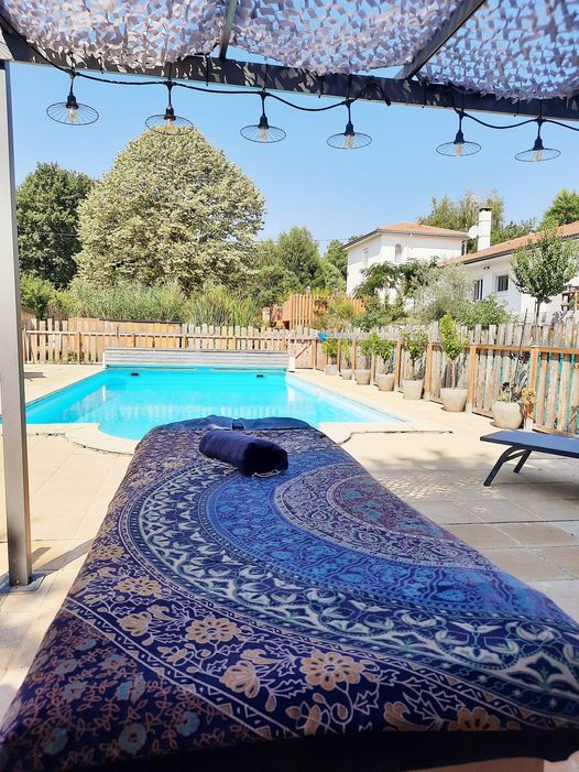 Un patio ensoleillé au bord de la piscine présente une chaise longue recouverte d'une serviette à motifs bleu et or, une serviette marine enroulée prête pour un massage relaxant, et une piscine claire entourée d'arbres et d'une maison blanche à l'arrière-plan.