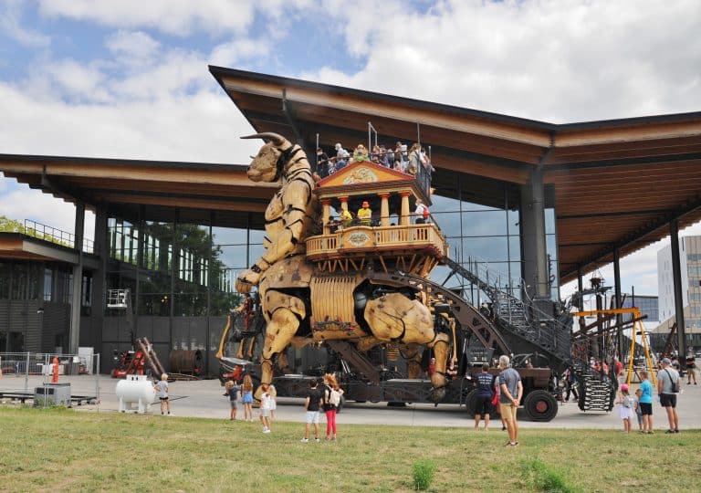 Un grand minotaure mécanique en bois sur lequel des personnes montent se déplace à l'extérieur d'un bâtiment moderne, sous le regard des badauds qui prennent des photos sur une pelouse, sous un ciel partiellement nuageux.