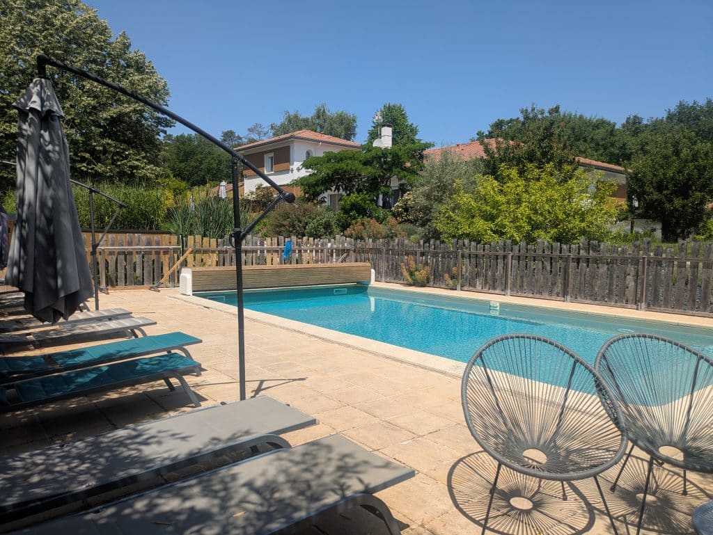 Une piscine extérieure ensoleillée entourée de chaises longues, d'un parasol fermé et de chaises grillagées, avec une clôture en bois, des arbres verts et des maisons en arrière-plan, sous un ciel bleu clair.