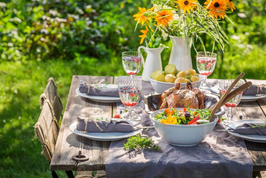 Une table rustique dressée pour un repas comprend du poulet rôti, un bol de salade, des verres décorés, des assiettes avec des serviettes, un pichet de pommes et un vase de fleurs jaunes sous la lumière du soleil dans un jardin.