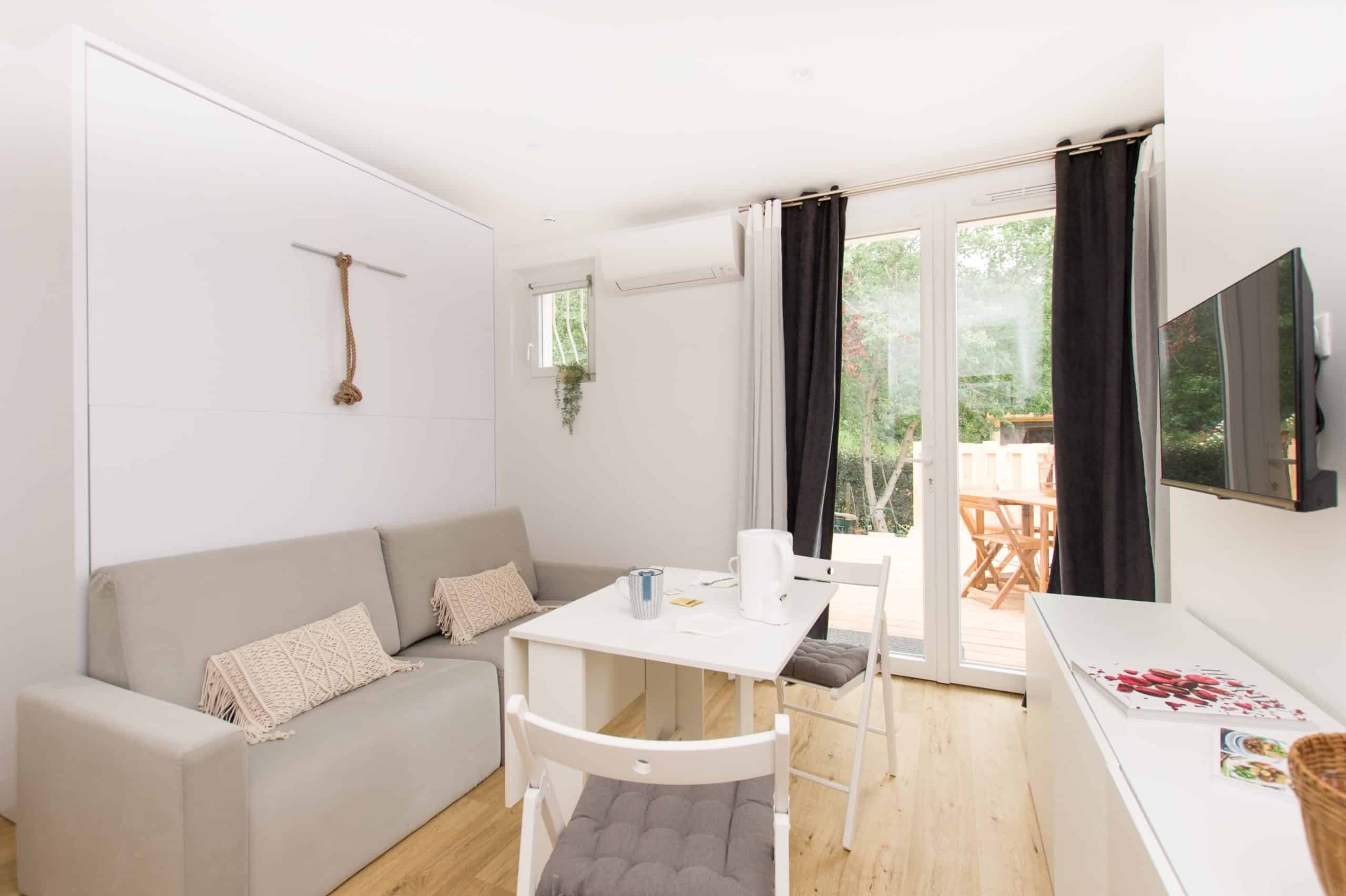 Ce gîte lumineux et moderne dispose d'un canapé gris, d'une petite table à manger avec deux chaises, d'une télévision murale et de grandes fenêtres avec des rideaux sombres donnant sur une terrasse en bois et la verdure à l'extérieur.