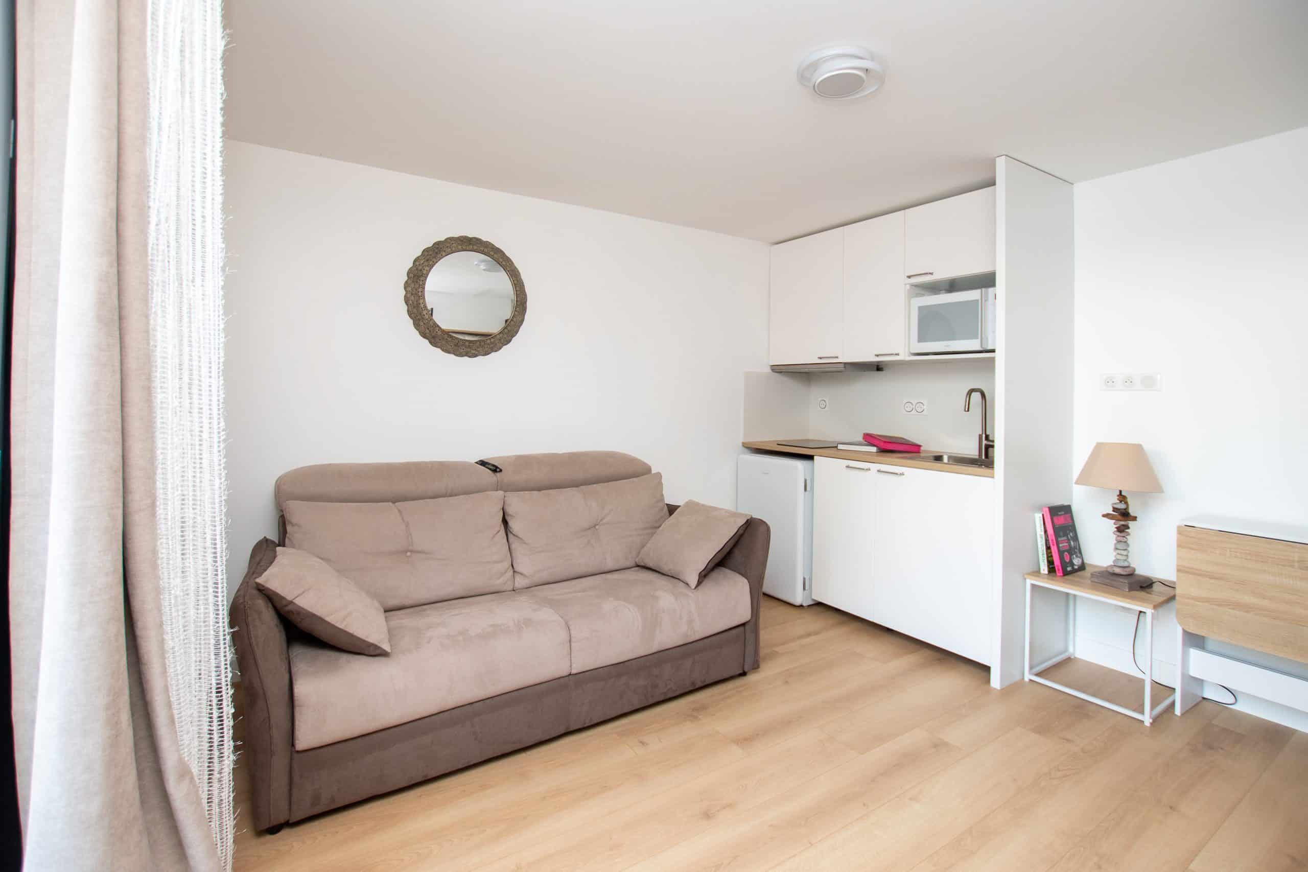 Ce petit studio lumineux, aménagé comme une chambre d'hôte, se compose d'un canapé beige, d'un miroir rond au mur, d'une kitchenette compacte avec des meubles blancs, d'une lampe sur une table d'appoint et d'un sol en bois clair.