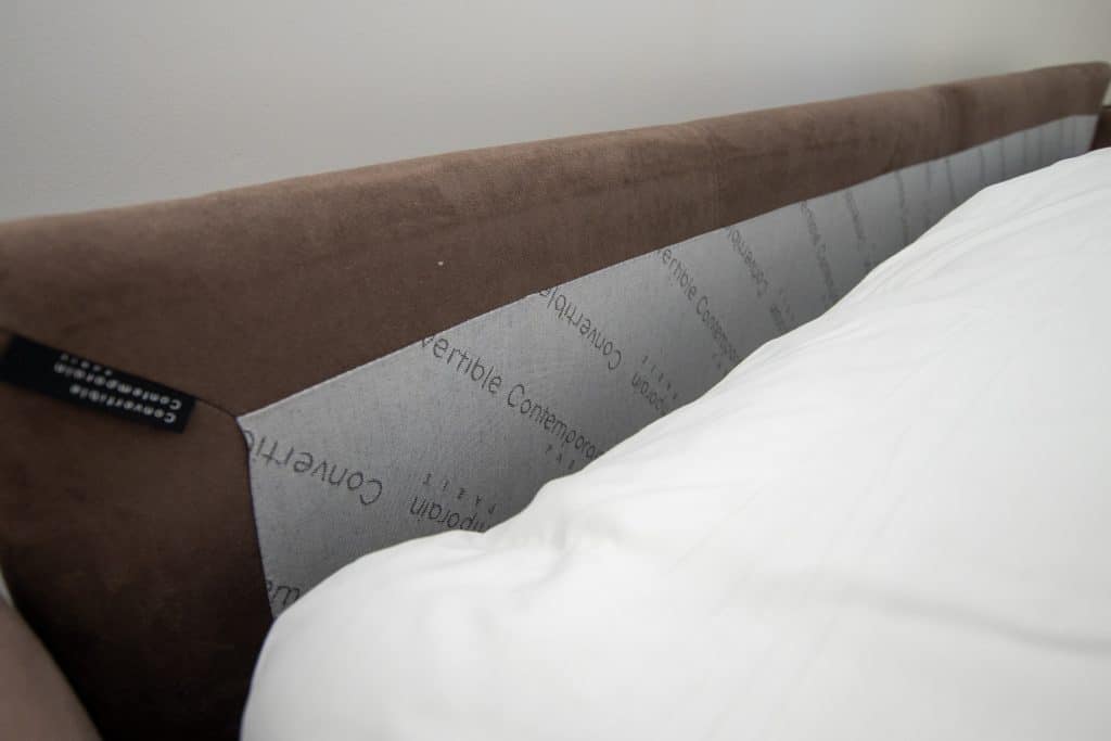 Gros plan sur une tête de lit capitonnée marron et grise, partiellement recouverte d'une couette blanche, avec une étiquette en tissu visible sur le côté gauche - parfaite pour une chambre d'hôte confortable ou pour être associée à un canapé convertible.