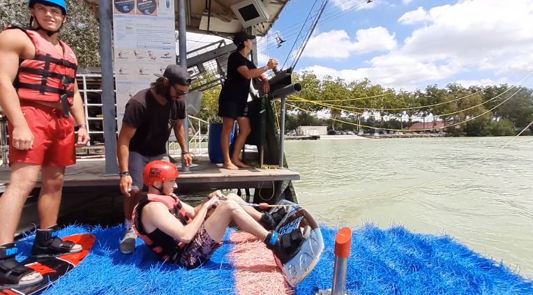 Groupe de personnes dans une station de wakeboard au bord d'un lac, près d'une chambre d'hôtes, avec une personne assise sur un tapis bleu se préparant à commencer, portant un casque et un gilet de sauvetage, tandis que d'autres personnes se tiennent à proximité sous un ciel partiellement nuageux.