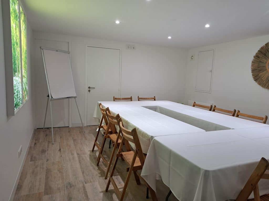 Une petite salle de réunion lumineuse avec un sol en bois, des tables en forme de U recouvertes de tissus blancs, des chaises en bois, un tableau à feuilles mobiles et un tableau sur le thème de la nature accroché à un mur. L'éclairage est assuré par des plafonniers.