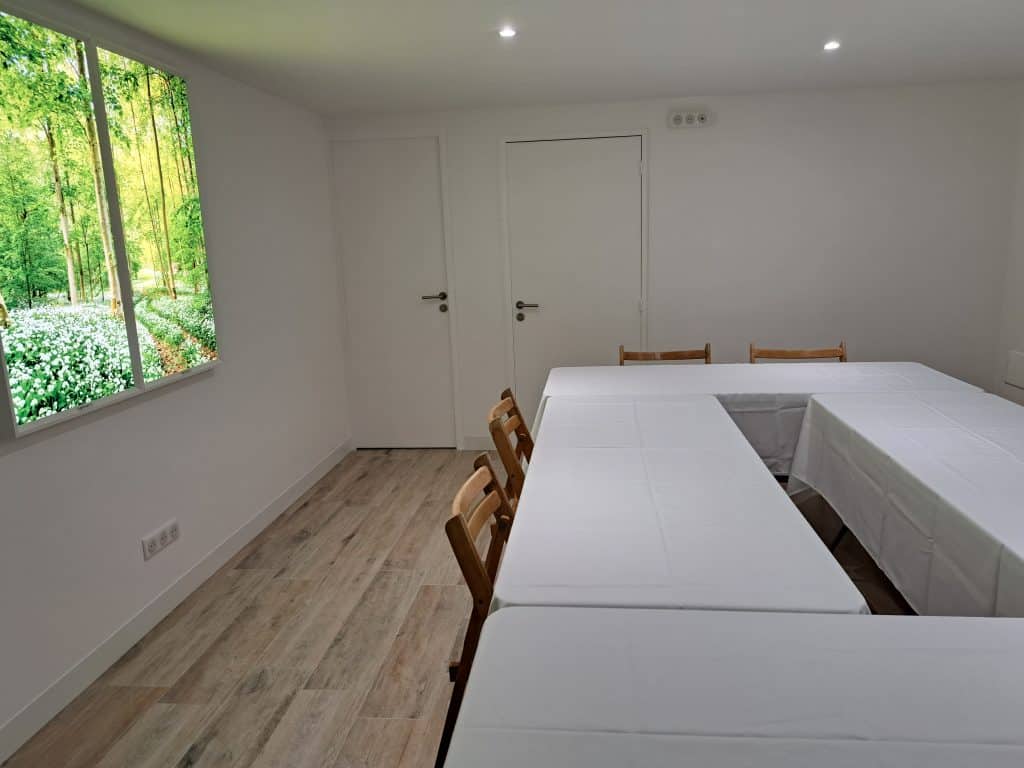 Une petite salle de réunion aux murs blancs, au sol en bois et à la table en forme de U recouverte de tissus blancs. Des chaises en bois sont disposées autour de la table. Un tableau lumineux donne une impression d'une grande fenêtre donnant sur une forêt verdoyante.