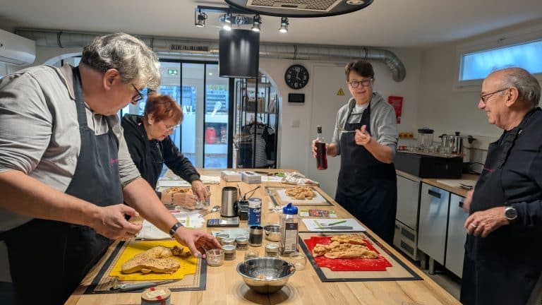 Quatre personnes en tablier préparent un foie gras. Les ingrédients et les ustensiles sont disposés sur le plan de travail. L'une d'entre elles fait un geste en tenant une bouteille, tandis qu'une autre saupoudre des épices.