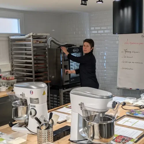 La chef Anouchka dans une cuisine se tourne face à la caméra alors qu'elle utilise un four commercial. La cuisine est équipée de mélangeurs, d'ustensiles et d'un tableau blanc avec des notes en français. Des plaques de cuisson sont empilées sur des étagères à l'arrière-plan.