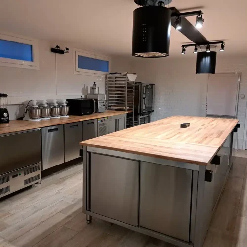 Un atelier de cuisine et pâtisserie spacieux et moderne avec un grand îlot en bois, des comptoirs en acier inoxydable, des mélangeurs, un micro-ondes, des étagères de boulangerie et des éclairages suspendus. Les murs sont blancs et le sol est en bois clair.
