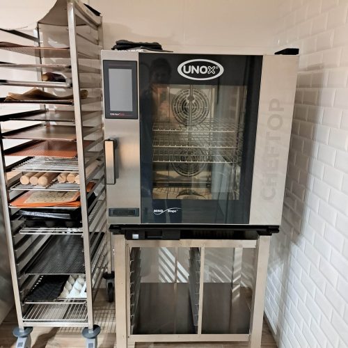 Un four commercial Unox ChefTop est posé sur un support métallique dans cet atelier de cuisine et pâtisserie, à côté d'une étagère remplie de plateaux de pâte et de produits de boulangerie dans une cuisine aux murs carrelés blancs.