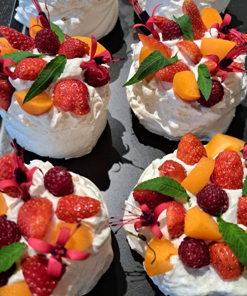 Mini pavlovas garnis de crème fouettée, de fraises, de framboises, de morceaux de fruits orange, de fleurs comestibles et de feuilles vertes, disposés sur un plateau noir.