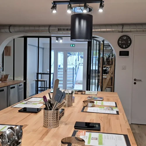 Une salle de classe moderne d'atelier cuisine et pâtisserie avec des postes de travail en bois équipés d'ustensiles, de fiches de recettes et d'outils de cuisine. Des appareils en acier inoxydable et une horloge sont visibles, et des portes en verre s'ouvrent sur un escalier à l'extérieur.