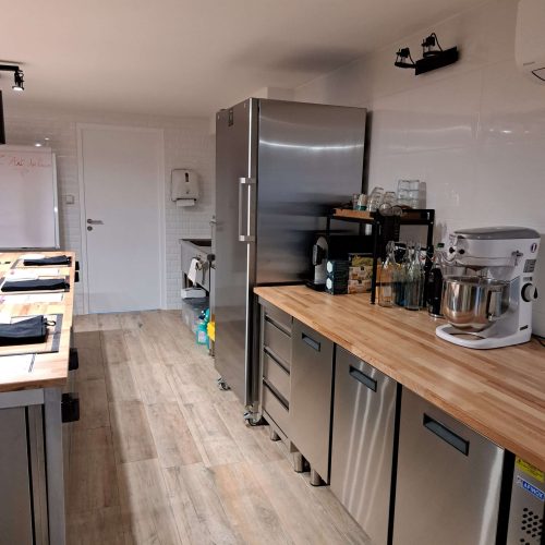 Un atelier moderne de cuisine et pâtisserie avec des appareils en acier inoxydable, des plans de travail en bois, un mixeur sur socle, de la verrerie, diverses bouteilles et des étagères. Le sol en bois et un grand réfrigérateur près du mur du fond complètent cette cuisine professionnelle.