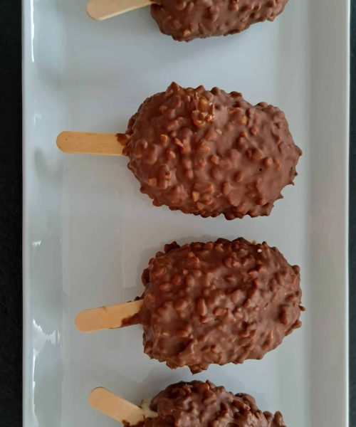 Quatre barres glacées enrobées de chocolat, à la texture croquante et noisetée, sont disposées en rang sur une assiette rectangulaire blanche, chacune sur un bâtonnet en bois.
