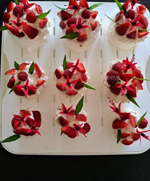 Huit petits desserts garnis de crème fouettée, de fraises en tranches et de feuilles vertes sont disposés proprement sur un plateau blanc. Le fond est sombre et quelques fraises sont placées sur le côté.
